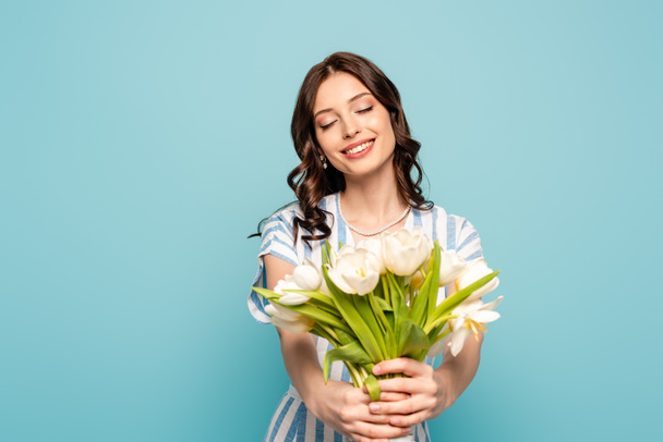gelukkig jong vrouw houden boeket van witte tulpen met gesloten ogen geïsoleerd op blauw - Foto, afbeelding