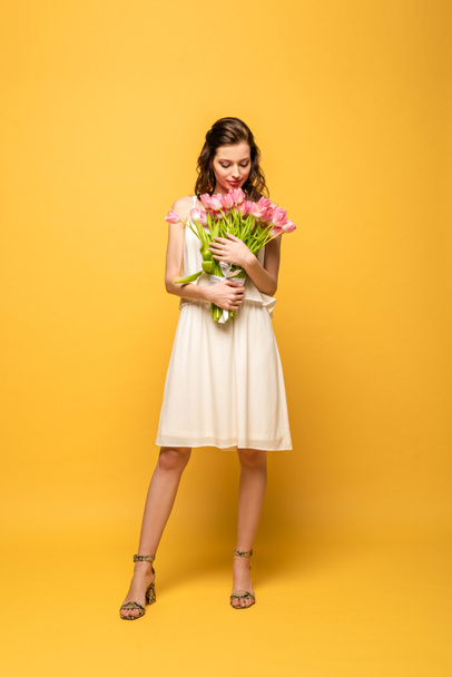 full length view of attractive young woman smiling while holding bouquet of pink tulips on yellow background - Photo, Image