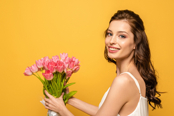 beautiful young woman smiling at camera while holding bouquet of pink tulips isolated on yellow - Foto, Imagem