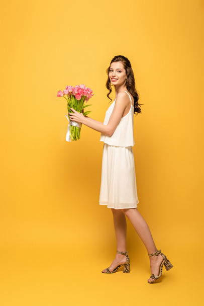 full length view of attractive young woman smiling at camera while holding bouquet of pink tulips on yellow background - Fotografie, Obrázek