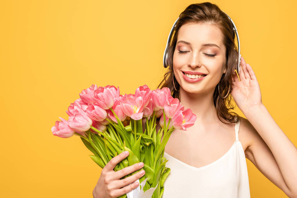 happy young woman holding bouquet of pink tulips while listening music in wireless headphones with closed eyes isolated on yellow - Φωτογραφία, εικόνα