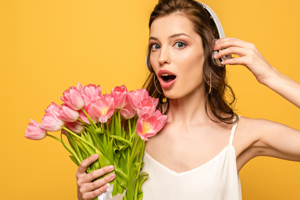 schockierte junge Frau mit drahtlosen Kopfhörern, die in die Kamera schaut, während sie einen Strauß rosa Tulpen auf gelb isoliert hält - Foto, Bild