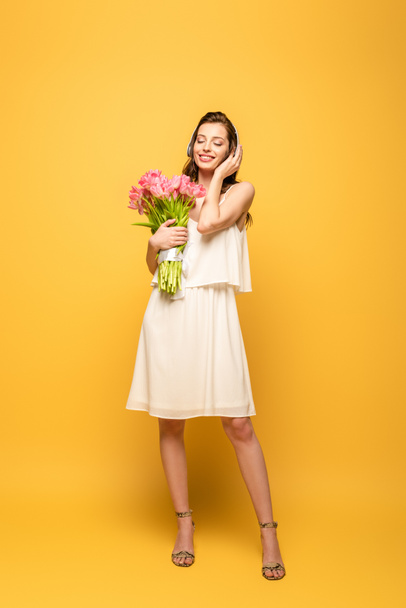 pełny widok długości szczęśliwy młoda kobieta w słuchawki bezprzewodowe gospodarstwa bukiet różowy tulipany na żółtym tle - Zdjęcie, obraz