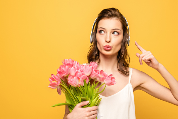 jeune femme réfléchie dans les écouteurs sans fil pointant avec le doigt tout en tenant bouquet de tulipes isolées sur jaune
 - Photo, image