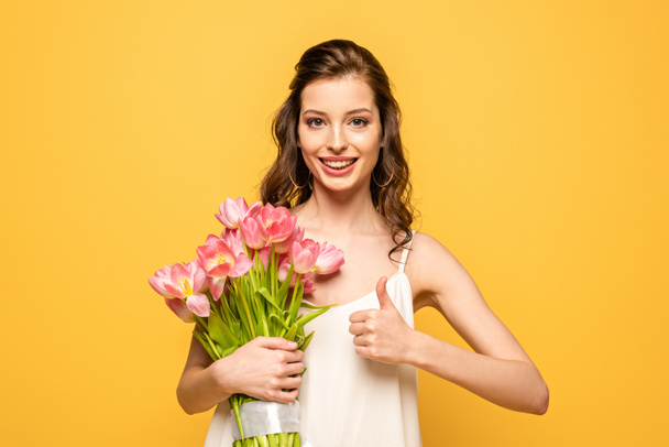 happy young woman holding bouquet of pink tulips while looking at camera and showing thumb up isolated on yellow - Valokuva, kuva
