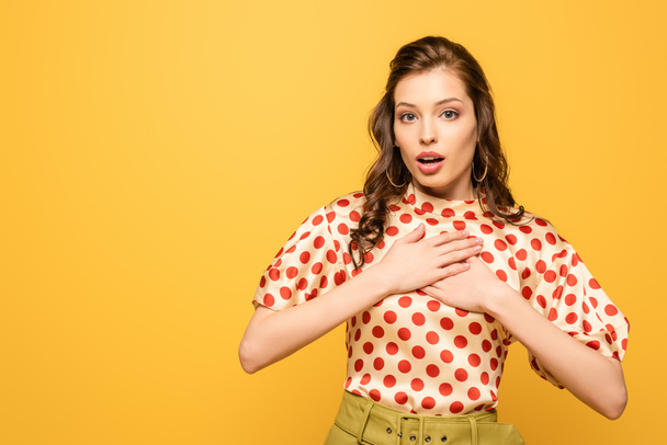 surprised young woman holding hands on chest while looking at camera isolated on yellow - Foto, imagen