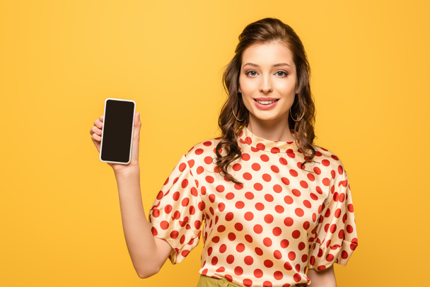 pretty young woman smiling at camera while showing smartphone with blank screen isolated on yellow - Photo, image