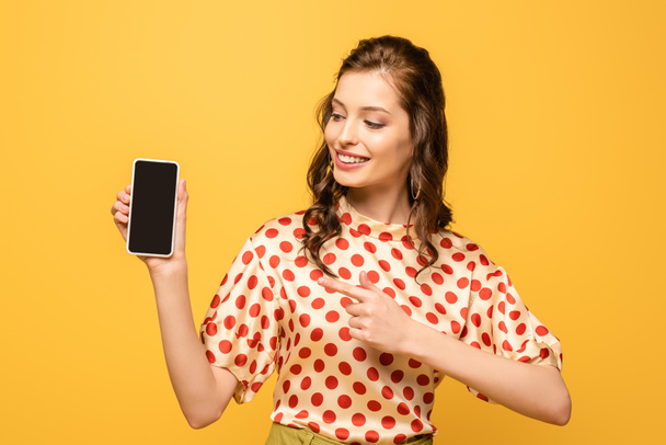 atrakcyjna młoda kobieta uśmiecha się wskazując palcem na smartfona z pustym ekranem odizolowanym na żółto - Zdjęcie, obraz
