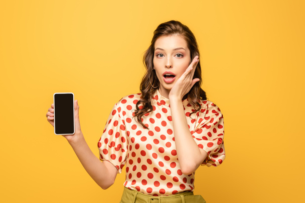 shocked young woman touching face while showing smartphone with blank screen isolated on yellow - Fotó, kép
