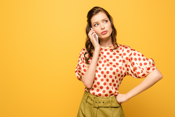 pensive young woman looking away while talking on smartphone isolated on yellow - Photo, Image