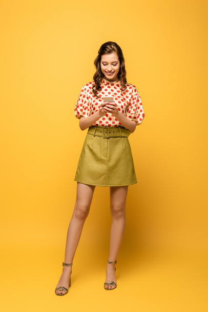 full length view of happy young woman chatting on smartphone on yellow background - Photo, Image