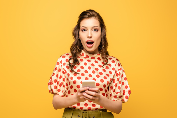 shocked young woman looking at camera while holding smartphone isolated on yellow - Zdjęcie, obraz