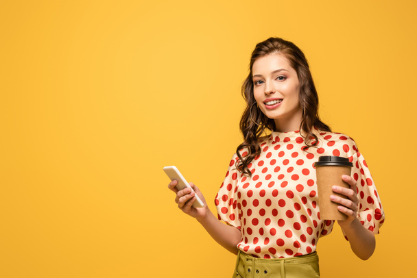 attractive young woman holding coffee to go while using smartphone and looking at camera isolated on yellow - Foto, immagini