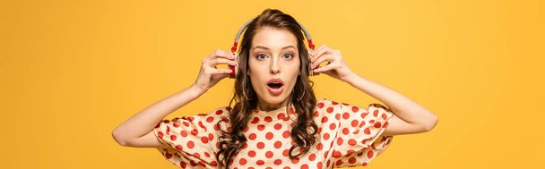 πανοραμική λήψη σοκαρισμένη νεαρή γυναίκα αγγίζει ασύρματα ακουστικά, ενώ κοιτάζοντας κάμερα που απομονώνονται σε κίτρινο - Φωτογραφία, εικόνα
