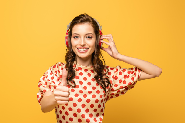 wesoła młoda kobieta w słuchawkach bezprzewodowych pokazuje kciuk w górę, uśmiechając się do kamery odizolowanej na żółto - Zdjęcie, obraz