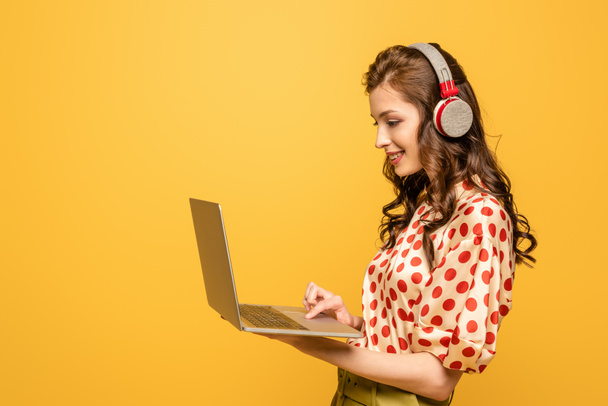 cheerful young woman in wireless headphones using laptop isolated on yellow - Zdjęcie, obraz