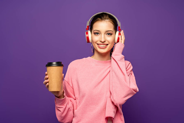 smiling girl holding coffee to go while listening music in wireless headphones isolated on purple - Fotografie, Obrázek