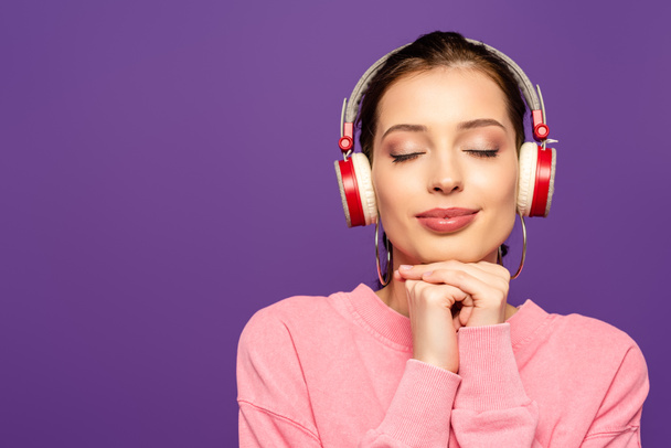 chica sonriente y soñadora escuchando música en auriculares inalámbricos con los ojos cerrados aislados en púrpura
 - Foto, Imagen