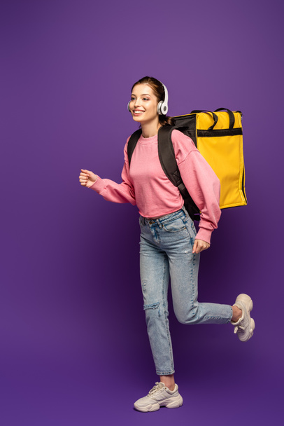 cheerful courier running in headphones while carrying thermo box on purple background - Foto, immagini