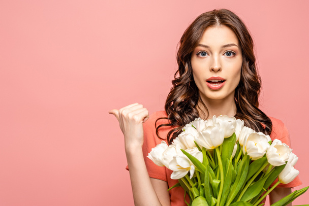 surprised young woman looking at camera and pointing with thumb while holding white tulips isolated on pink - Photo, Image