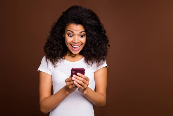 Портрет враженого африканського жіночого блогера, який користується мобільним телефоном, отримує повідомлення соціальної мережі, що виглядає кричущим, одягненим у білу футболку, ізольовану на коричневому тлі. - Фото, зображення