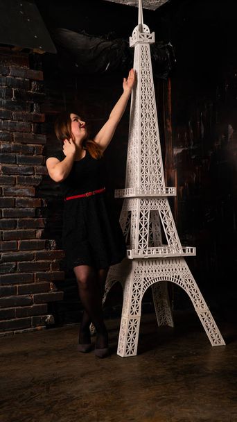 atrakcyjna kobieta w czarnej sukience pozowanie w pobliżu dekoracyjnej wieży Eiffla  - Zdjęcie, obraz