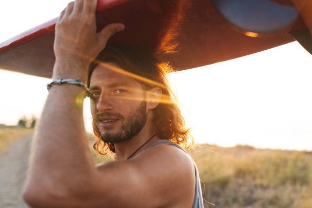 Photo of pleased unshaven man looking at camera holding surfboard - Photo, image