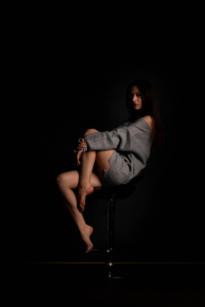 Sexy young in beautiful Gray dress with sleeves  posing in the studio on the chair On a black background  - 写真・画像