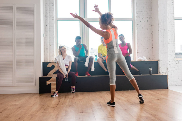 Enfoque selectivo de la mujer realizando movimientos con bailarinas de zumba multiculturales descansando en banco en estudio de danza
 - Foto, imagen