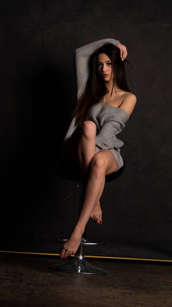 sexy jong in mooi grijs jurk met mouwen poseren in de studio op de stoel op een zwarte achtergrond  - Foto, afbeelding