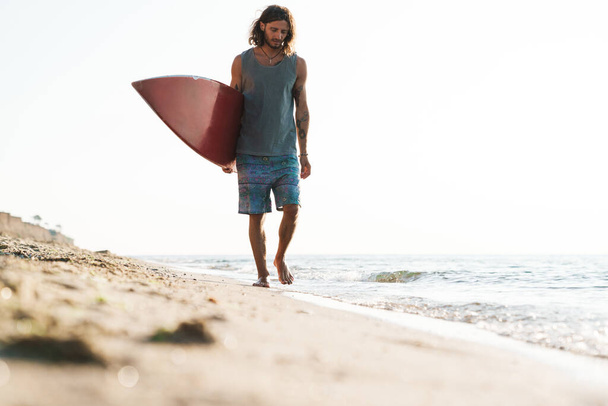 Photo of unshaven focused man holding surfboard while walking - Foto, imagen