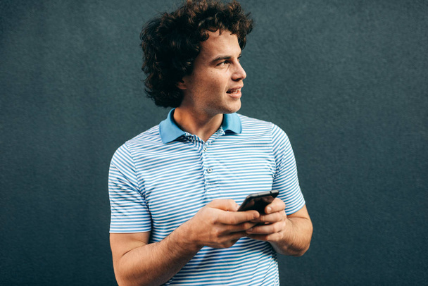 Young smart man browsing on the web on mobile phone. Handsome male with curly hair resting outside making a call on his smart phone in the city street. Lifestyle, people - Zdjęcie, obraz