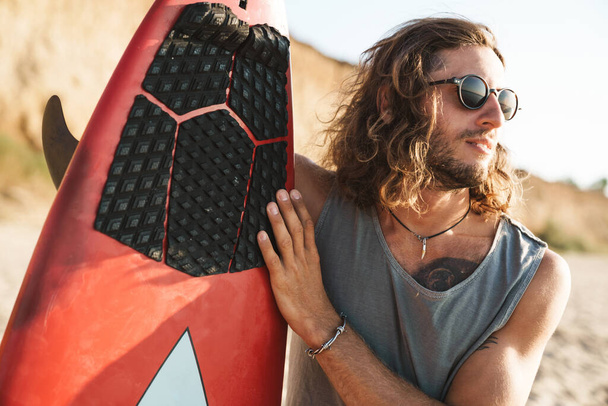 Photo of handsome confident man holding surfboard and looking forward - Фото, изображение