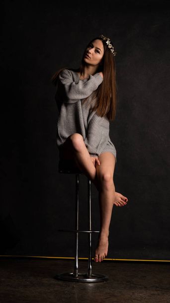 Sexy young in beautiful Gray dress with sleeves  posing in the studio on the chair On a black background  - Фото, зображення