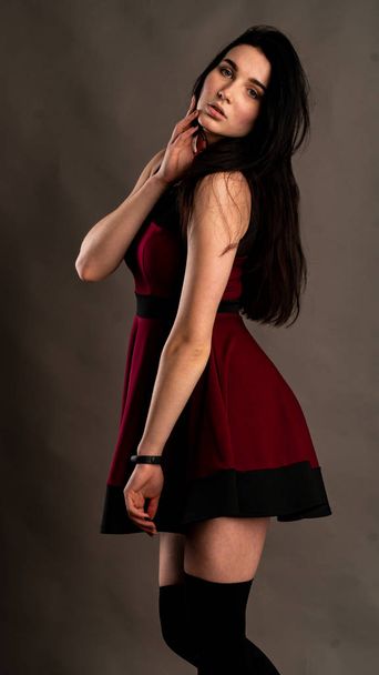Pretty young sexy model female with dark hair in red dress posing in dark studio. - Фото, изображение