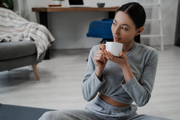 Gyönyörű fiatal nő sport egyenruhában otthon kávét iszik reggelire reggeli után gyakorlatok ülnek a padlón. - Fotó, kép
