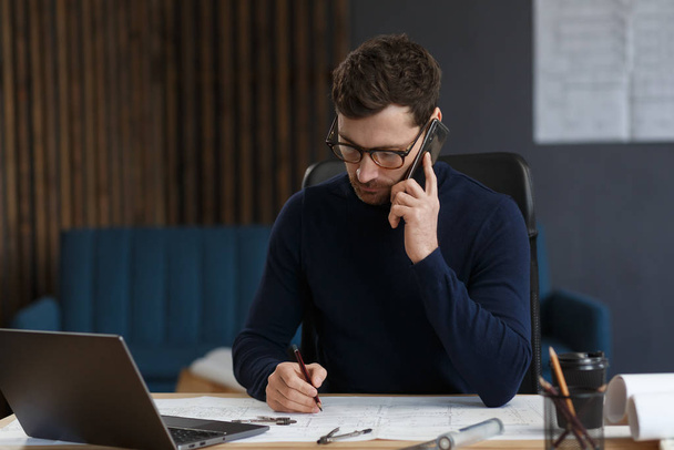 Architect talking on the phone in office. Confident businessman negotiates with business partners. Business portrait of handsome bearded man wearing eyeglasses sitting at workplace. Business concept - Фото, зображення