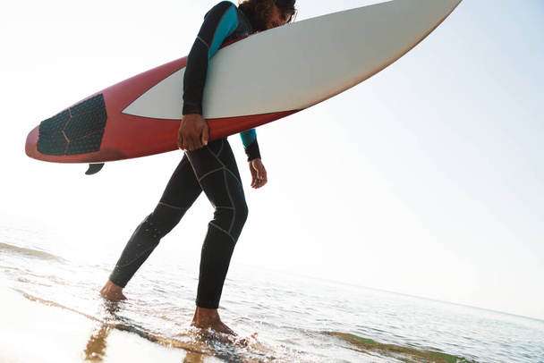 Foto de un joven caucásico sosteniendo la tabla de surf mientras camina
 - Foto, imagen