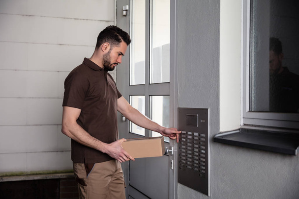 Side View Of Man Holding Cardboard Box Pressing Button Of Intercom To Enter Home - 写真・画像