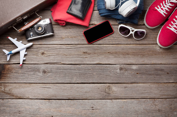 Suitcase, camera, clothes and travel accessories. Sneakers, jeans, smartphone, headphones and sunglasses. Tourist and vacation things on wooden background with space for your text. Top view flat lay - 写真・画像