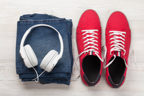 Clothing and accessories. Sneakers, jeans and headphones. Urban outfit for everyday or travel vacation on wooden background. Top view flat lay - Photo, image