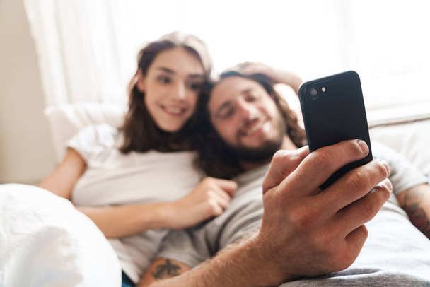 Loving couple indoors at home lies using mobile phone together. - 写真・画像