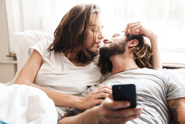 Loving couple indoors at home lies using mobile phone together. - Fotoğraf, Görsel