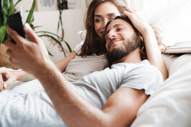 Loving couple indoors at home lies using mobile phone together. - Foto, Imagen