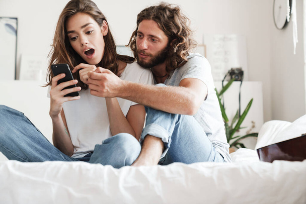 Couple indoors at home sit on bed using mobile phone. - Photo, Image