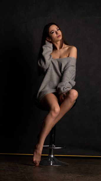 Sexy joven con hermoso vestido gris con mangas que posan en el estudio en la silla En un fondo negro.  - Foto, imagen