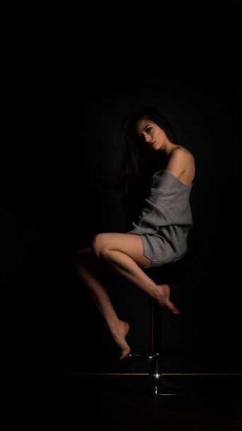 Sexy junge in schönen grauen Kleid mit Ärmeln posiert im Studio auf dem Stuhl Auf einem schwarzen Hintergrund  - Foto, Bild