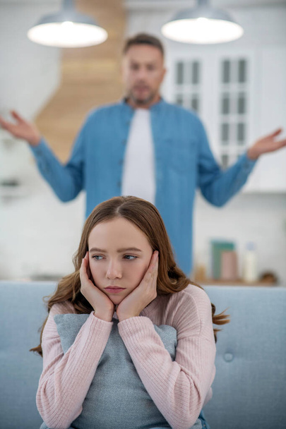 Dad standing behind a sad daughter sitting on sofa. - Фото, изображение