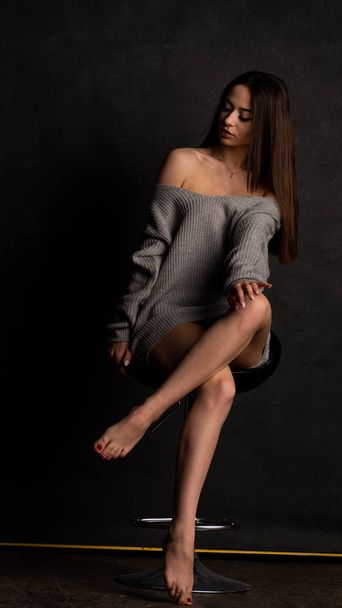 Jeune sexy en belle robe grise avec des manches posant dans le studio sur la chaise Sur un fond noir  - Photo, image