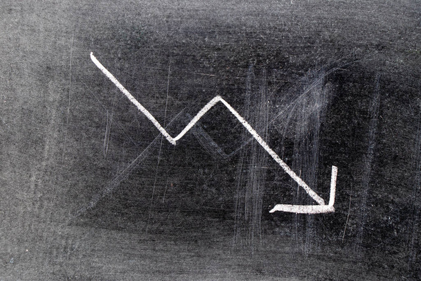 Craie blanche dessin à la main en forme de flèche vers le bas sur fond de tableau noir (Concept de déclin des stocks, tendance à la baisse des entreprises, économie) - Photo, image